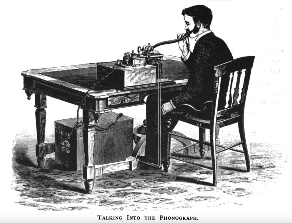 A man speaking into a tube connected to a late 19th century phonograph
